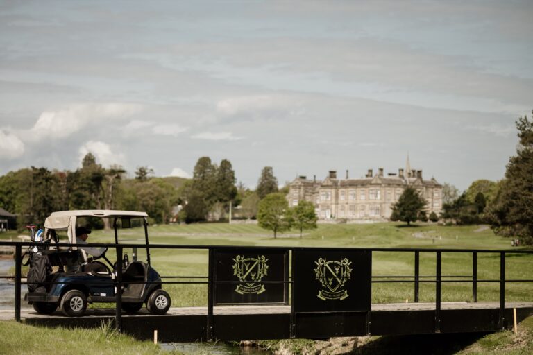 A golf buggy crossing a bridge on the course, with the hall in the distance, Matfen Hall