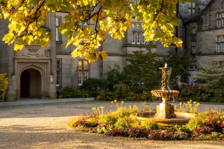 The stone fountain on the forecourt in autumn light, with yellow leaves overhead and the Gothic entrance arch behind, Matfen Hall