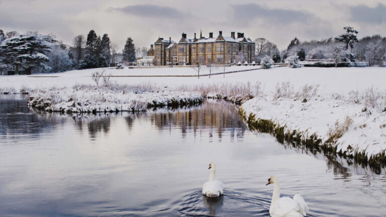 The hall across a snow-covered park in winter, with swans on the river in the foreground, Matfen Hall