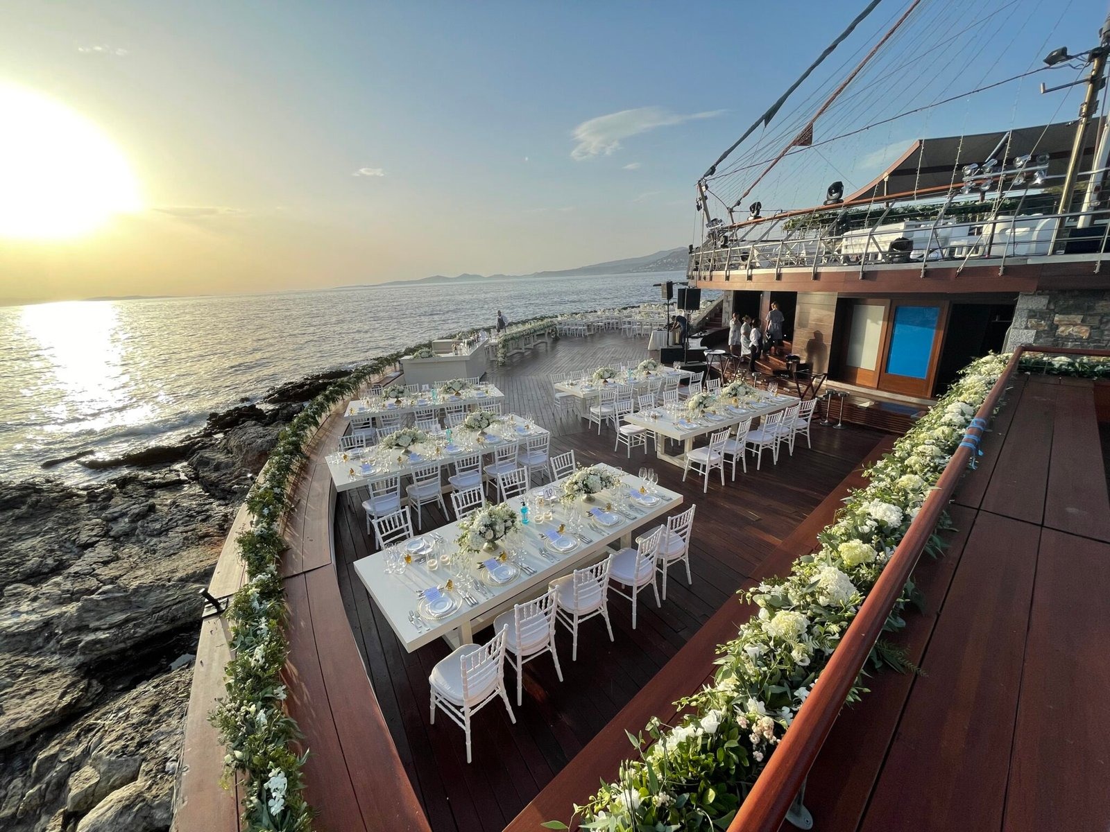 Dinner tables on a wooden terrace above the water, edged with greenery, Mathioudakis Exclusive Events