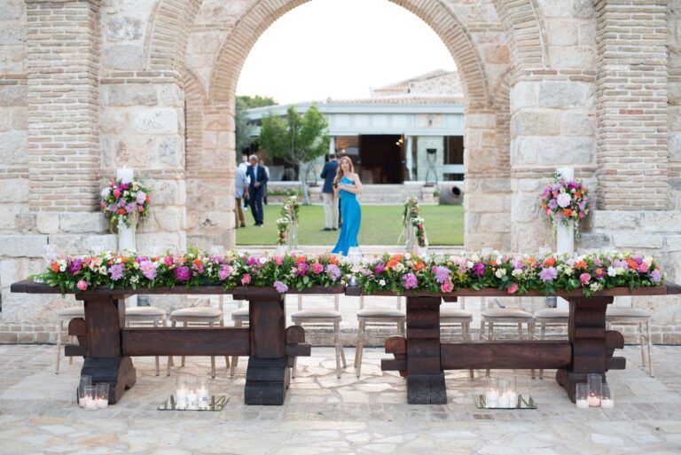 Long wooden table with a low flower runner before a stone arch, Mathioudakis Exclusive Events