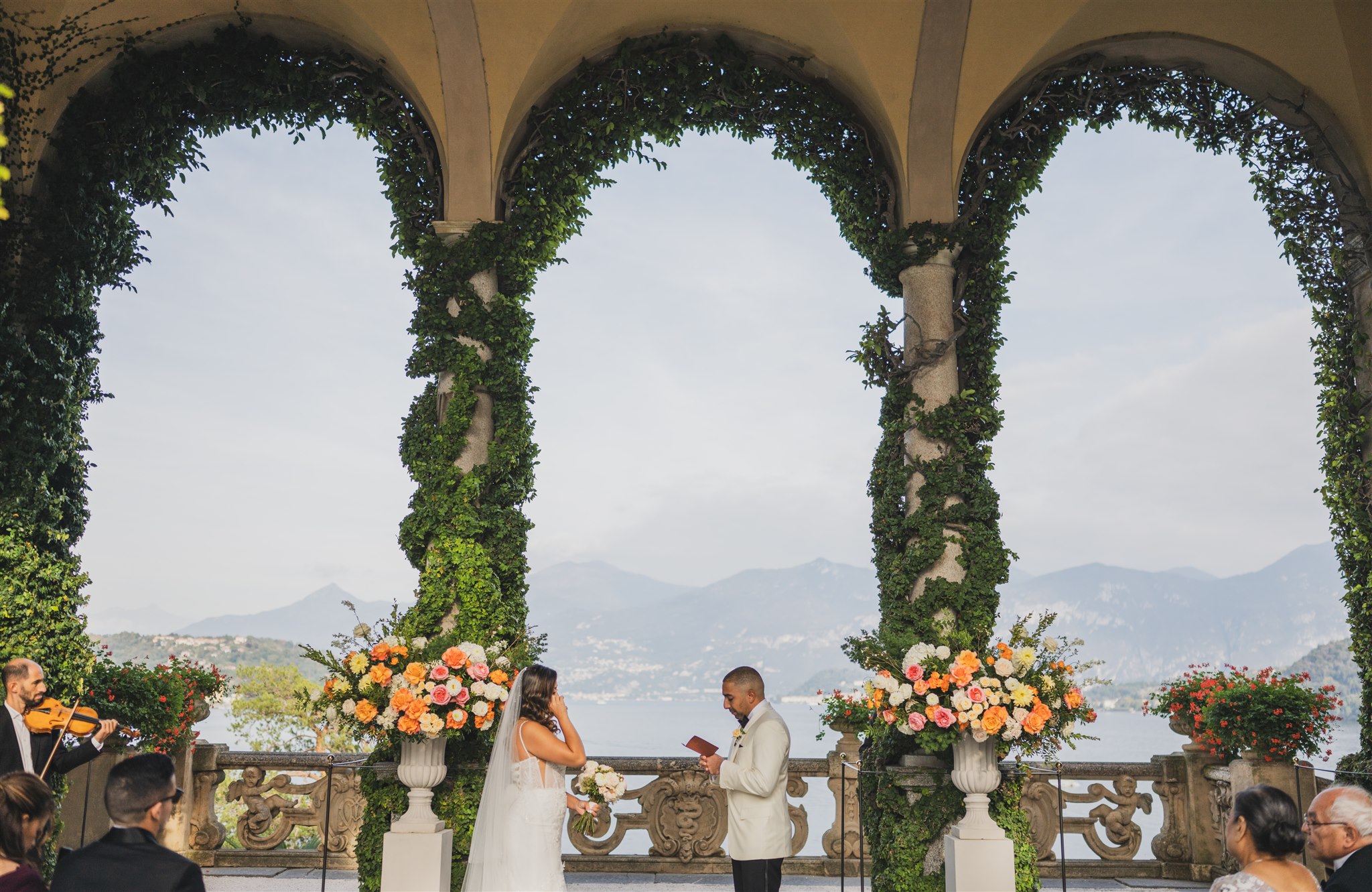 A ceremony under the ivy-covered loggia at Villa del Balbianello, the lake and mountains framed by the arches, Matteo Cuzzola Photography