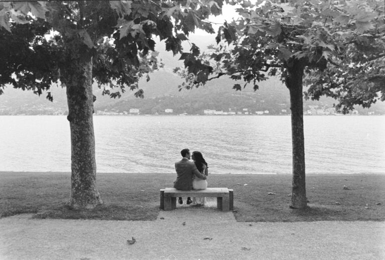 A couple on a bench between two trees on the shore of Lake Como, shot on black and white film, Matteo Cuzzola Photography