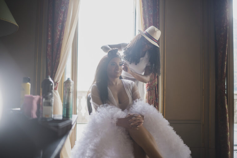 A bride getting ready by the window, a friend in a straw hat helping, from his Villa Erba page, Matteo Cuzzola Photography