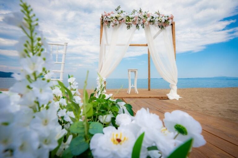 A wooden arch with white drapes and white flowers above a beach platform, Mazal Weddings