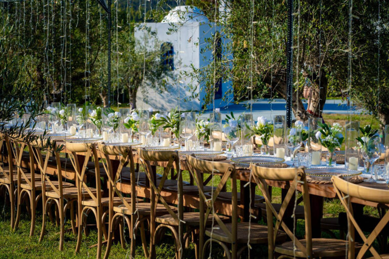 Long reception table beneath the olive trees with the chapel behind, Mazal Weddings