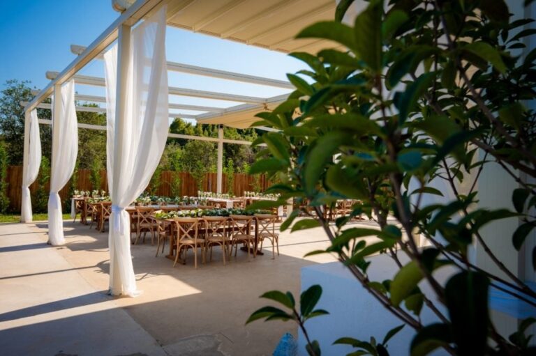 A covered terrace with long wooden tables and white drapes by day, Mazal Weddings
