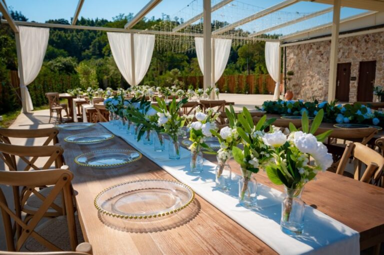 Long wooden tables with glass chargers and white flowers under the pergola, Mazal Weddings