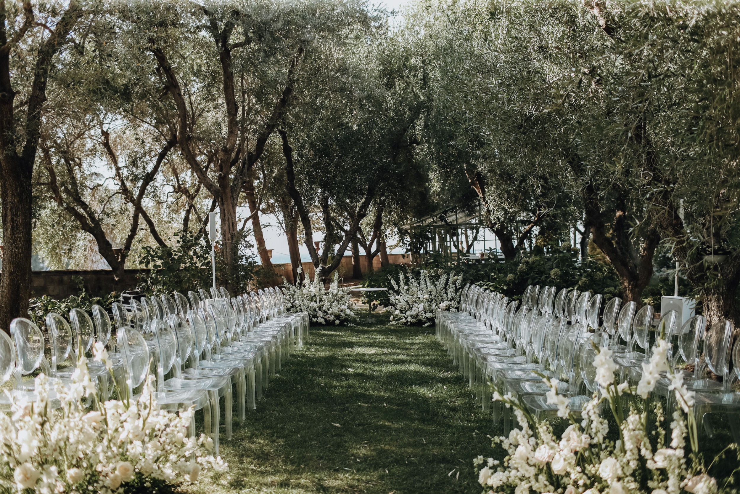 Rows of clear chairs either side of a grass aisle in an olive grove, banks of white flowers along the edges, Med inStyle