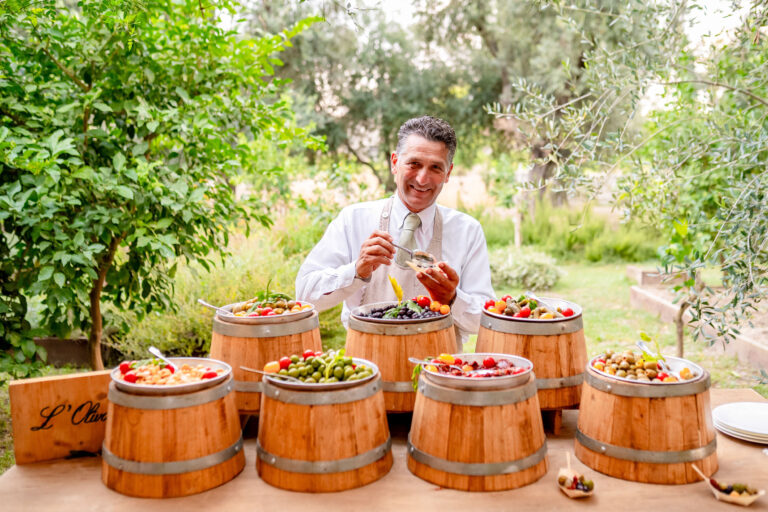 A waiter serving olives, tomatoes and pickles from small wooden barrels at an antipasti station in a citrus garden, Med inStyle