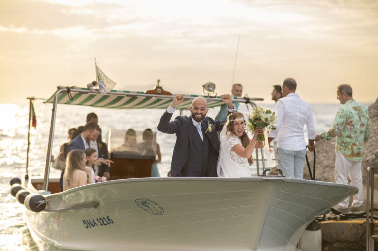 The bride and groom arriving by wooden boat at sunset, guests on board and on the jetty, Med inStyle