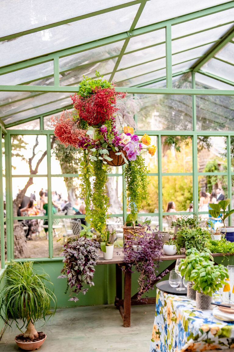A green-framed glass pavilion with a hanging installation of red flowers and trailing greenery, potted herbs on the table below, Med inStyle