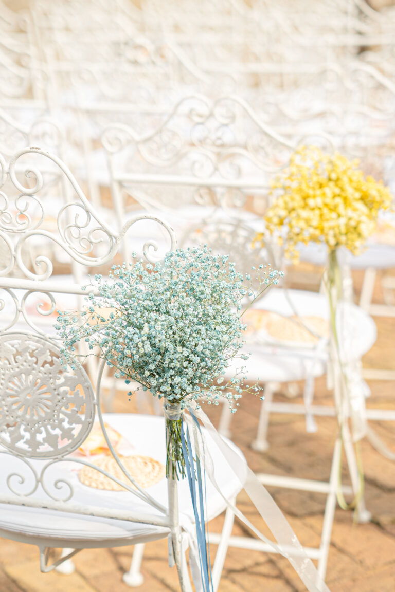 Small blue and yellow gypsophila posies tied to white wrought-iron ceremony chairs, Med inStyle