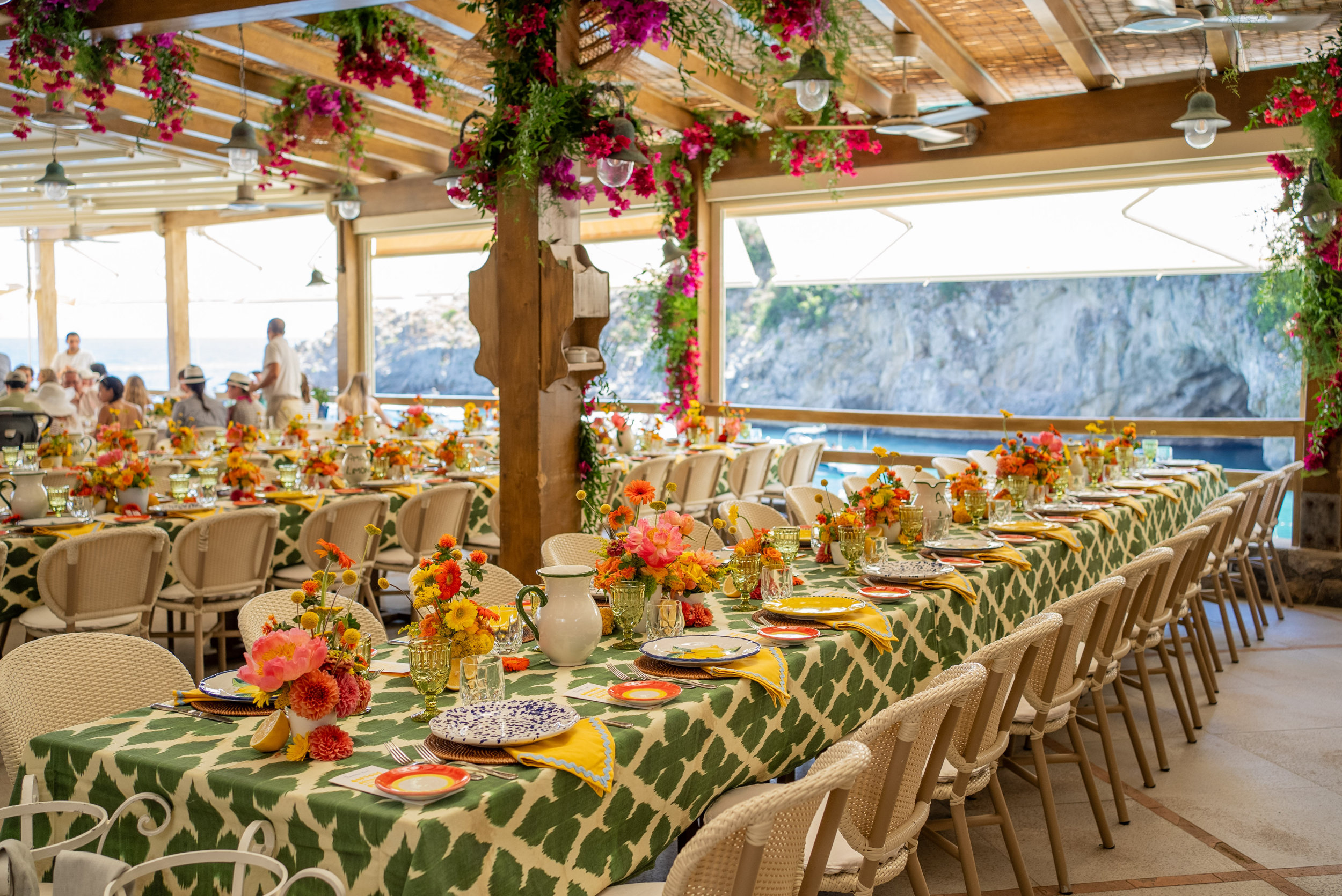 Long tables under a bougainvillea pergola, green patterned cloths, orange flowers and painted plates, the sea and cliffs beyond, Med inStyle