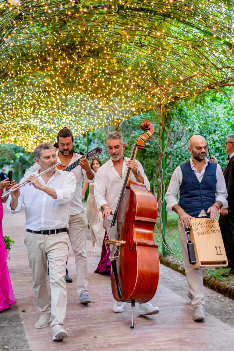 Musicians with flute, guitar, double bass and cajon walking beneath a pergola strung with fairy lights, Med inStyle