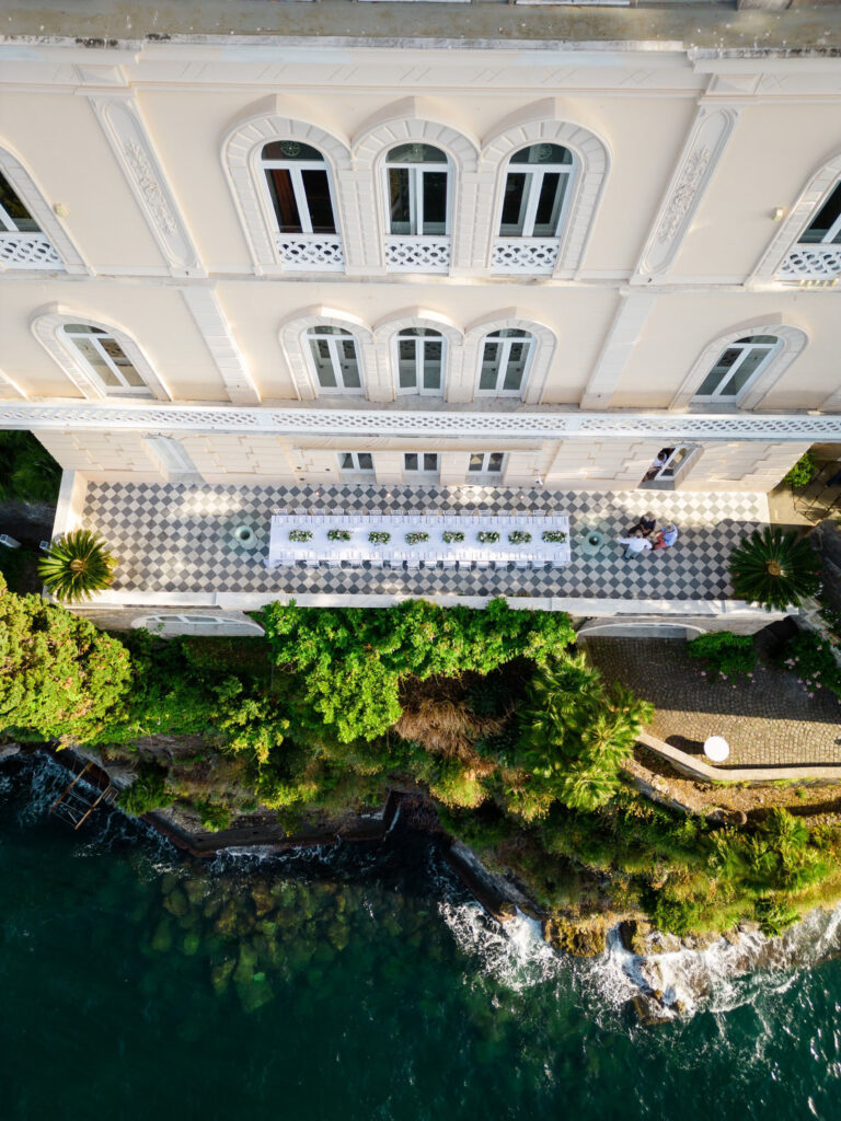Aerial view of a single long table on a chequered terrace in front of a white hotel facade, the cliff edge and sea below, Med inStyle
