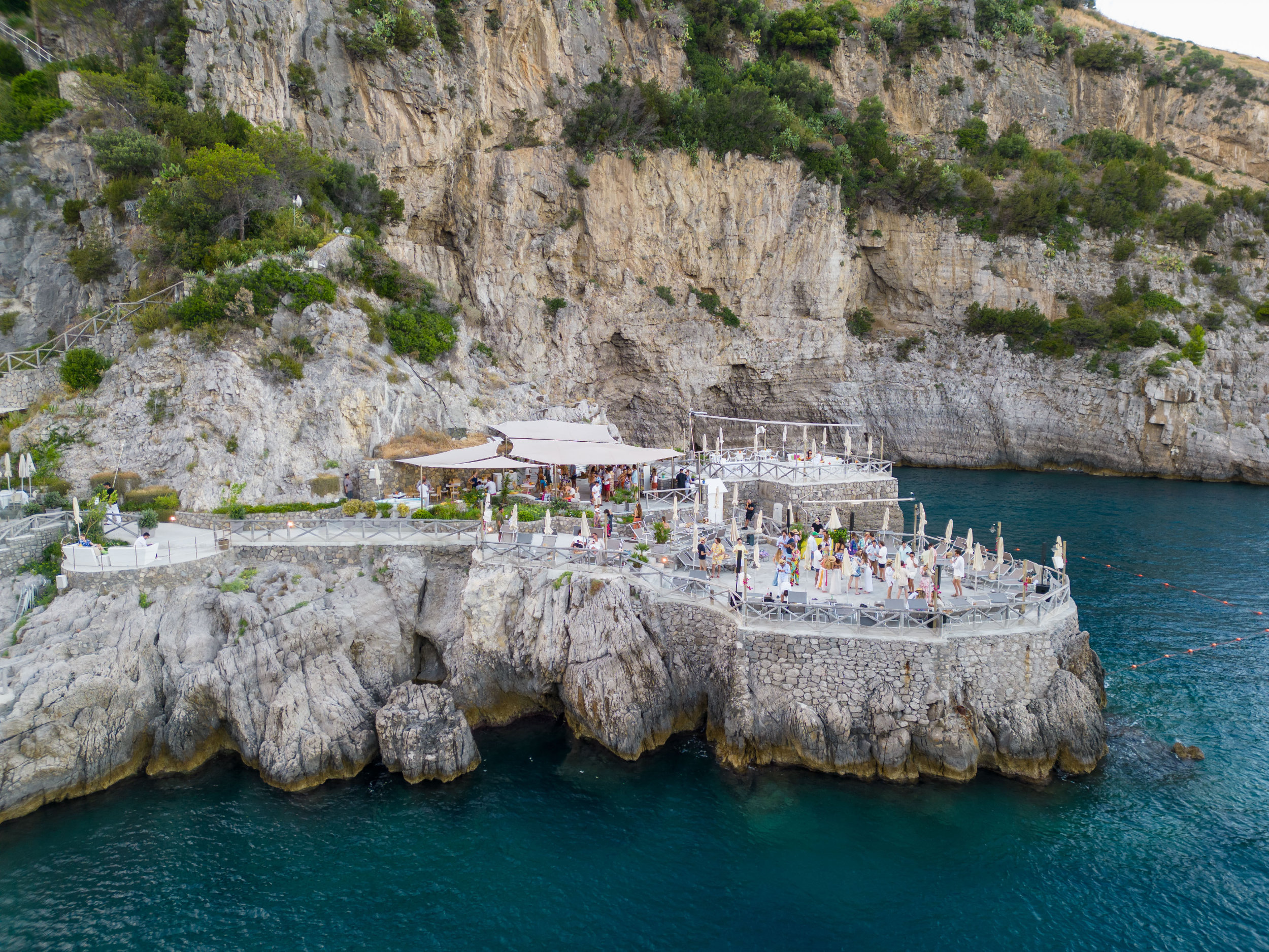Aerial view of a reception terrace built into the cliffs above a sheltered cove, guests gathered at the water's edge, Amalfi Coast, Med inStyle