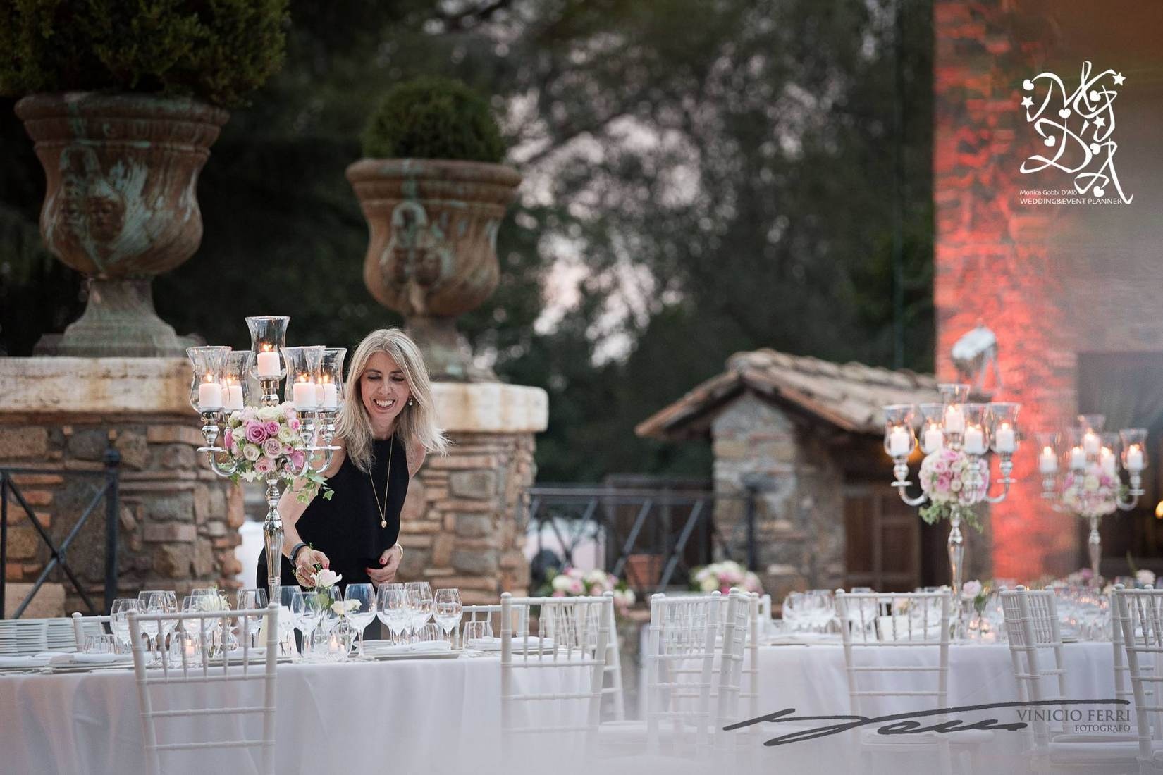Monica Gobbi D'Alò dresses a silver candelabra centrepiece on a long reception table at dusk, stone garden urns behind her, MGDA Events