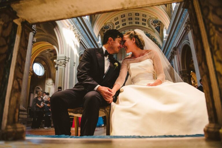 A bride and groom sit forehead to forehead inside a domed, frescoed basilica nave, MGDA Events