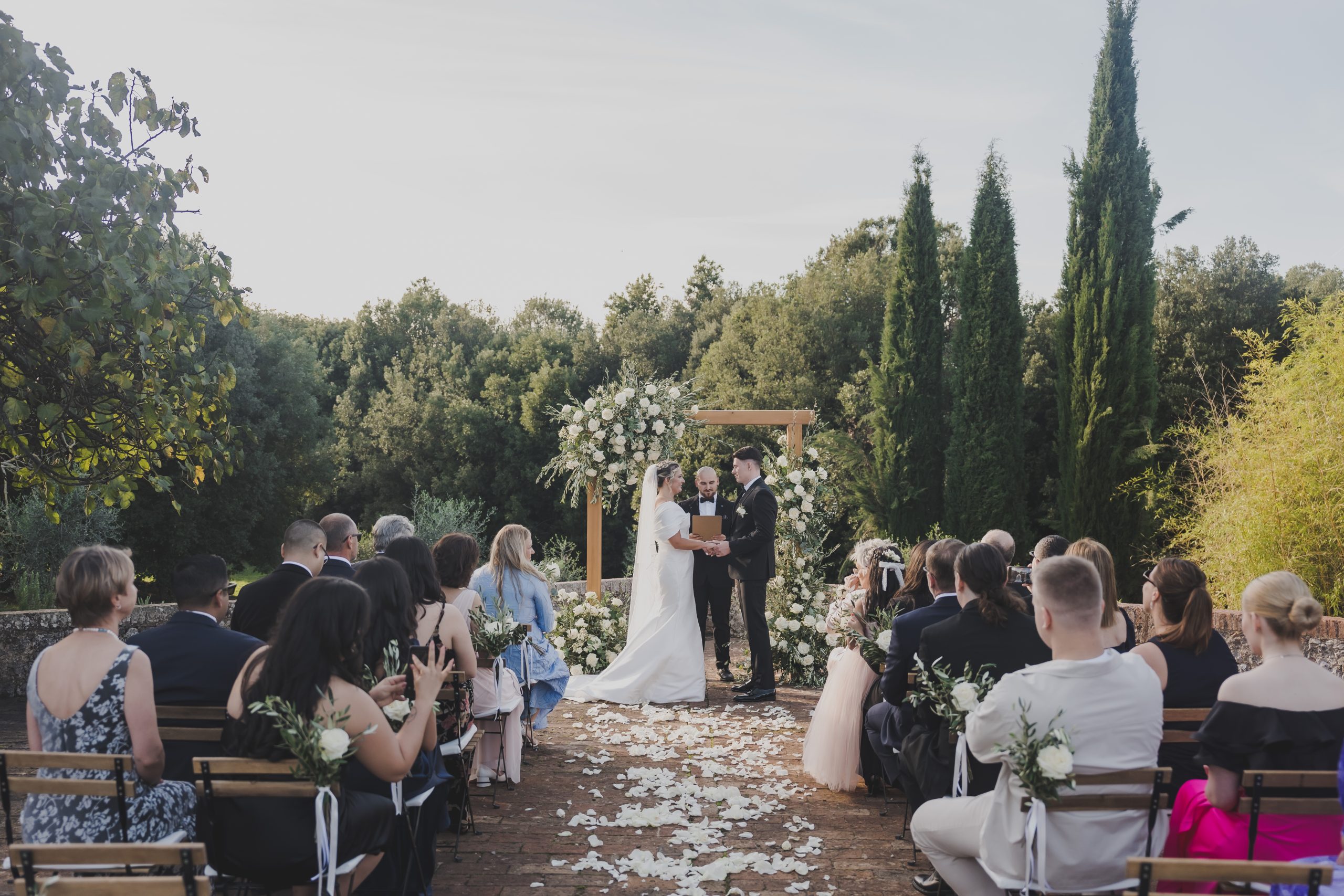 An outdoor ceremony at Borgo Gallinaio, guests seated on a petal-strewn terrace beneath tall cypresses, Michele Monasta Photography