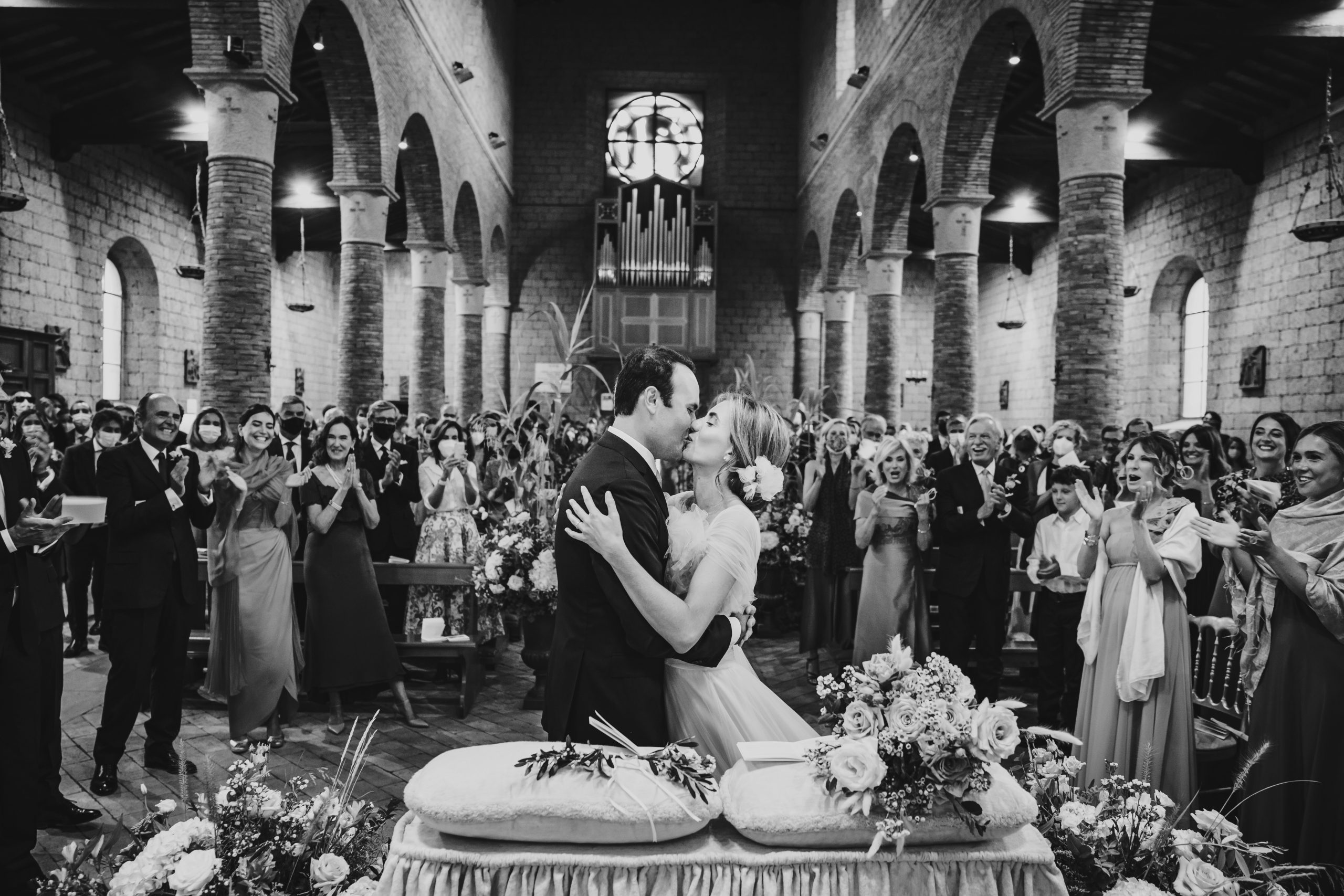 A first kiss as husband and wife in a crowded stone church, guests applauding, on the first of two days of celebration based at Borgo Carige in the Maremma, Michele Monasta Photography