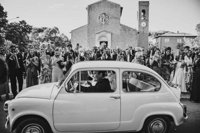 The newlyweds drive off in a vintage Fiat 600 while guests line the church piazza with their phones raised, Michele Monasta Photography