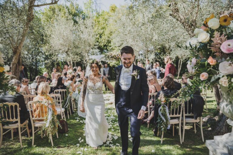 The couple walk back down an aisle lined with flowers among the olive trees at Castello di Bibbione, Michele Monasta Photography