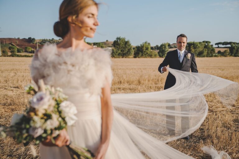The groom lifts the bride's long veil in a harvested wheat field in the Tuscan Maremma, Michele Monasta Photography