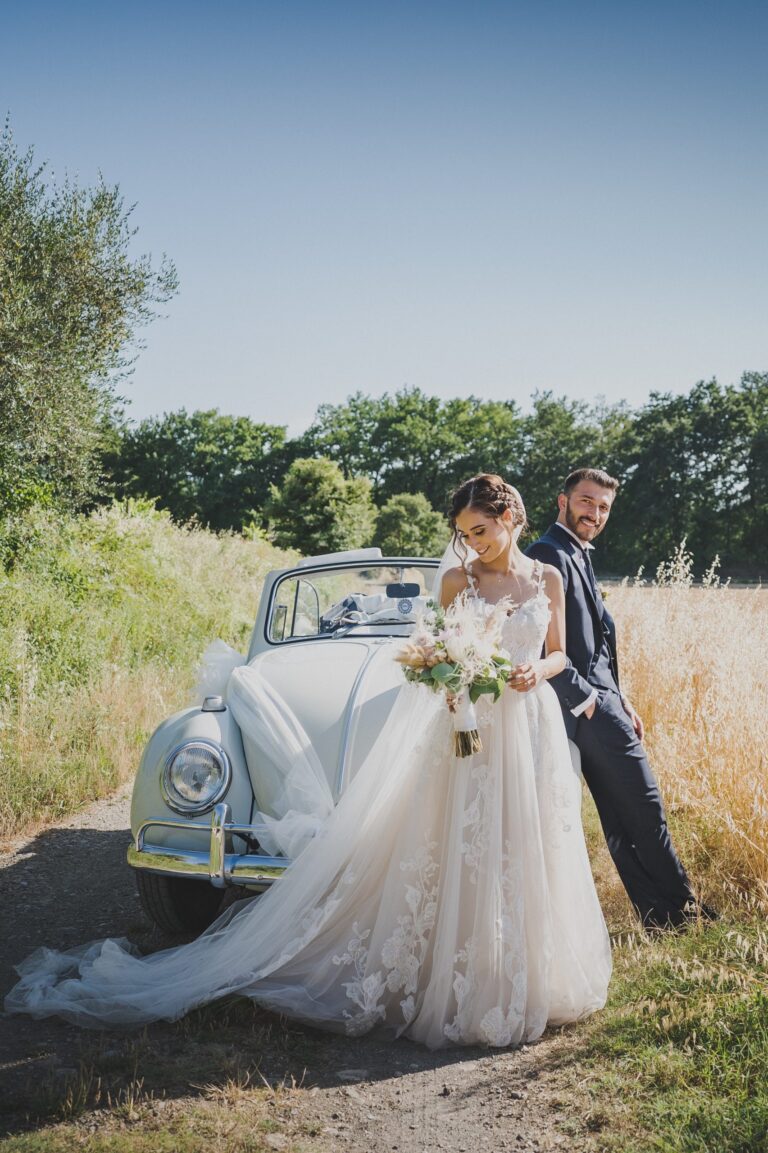 The couple beside a vintage convertible in a summer field, on the day they married at the Pieve di Borgo San Lorenzo in the Mugello, Michele Monasta Photography