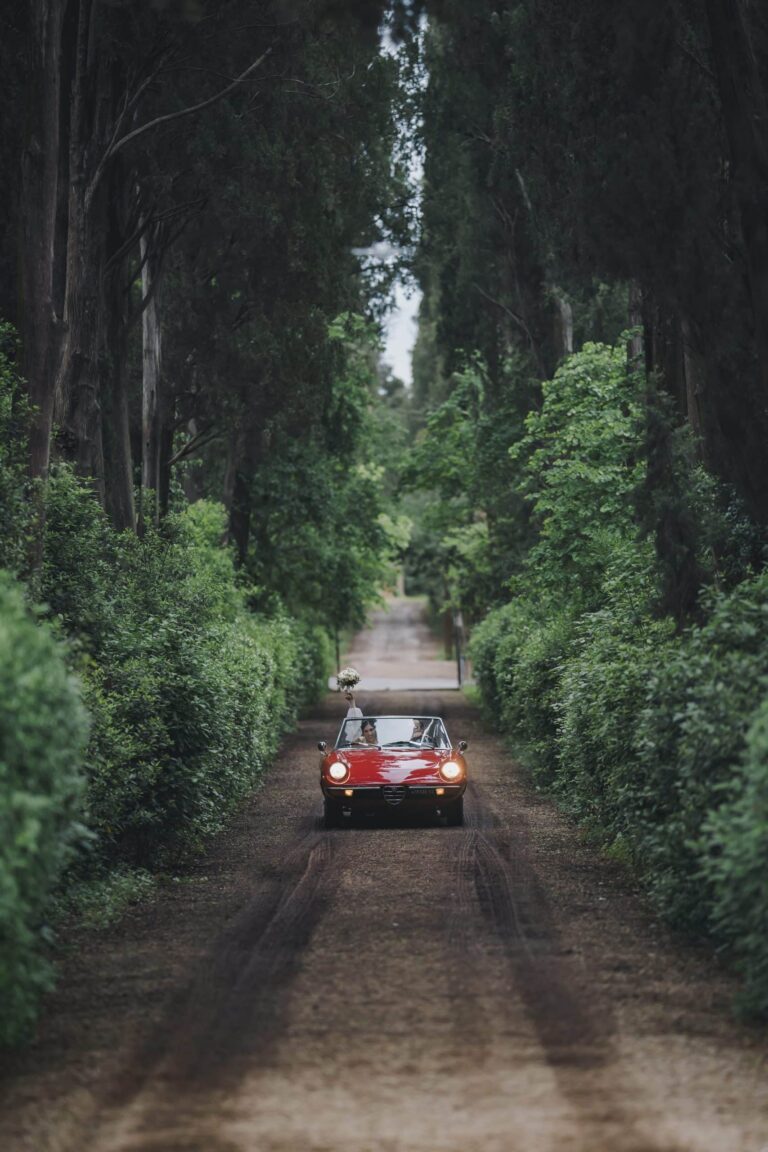 A red convertible comes down a cypress avenue, from a Florence wedding with its reception at Villa Le Corti in Chianti, Michele Monasta Photography
