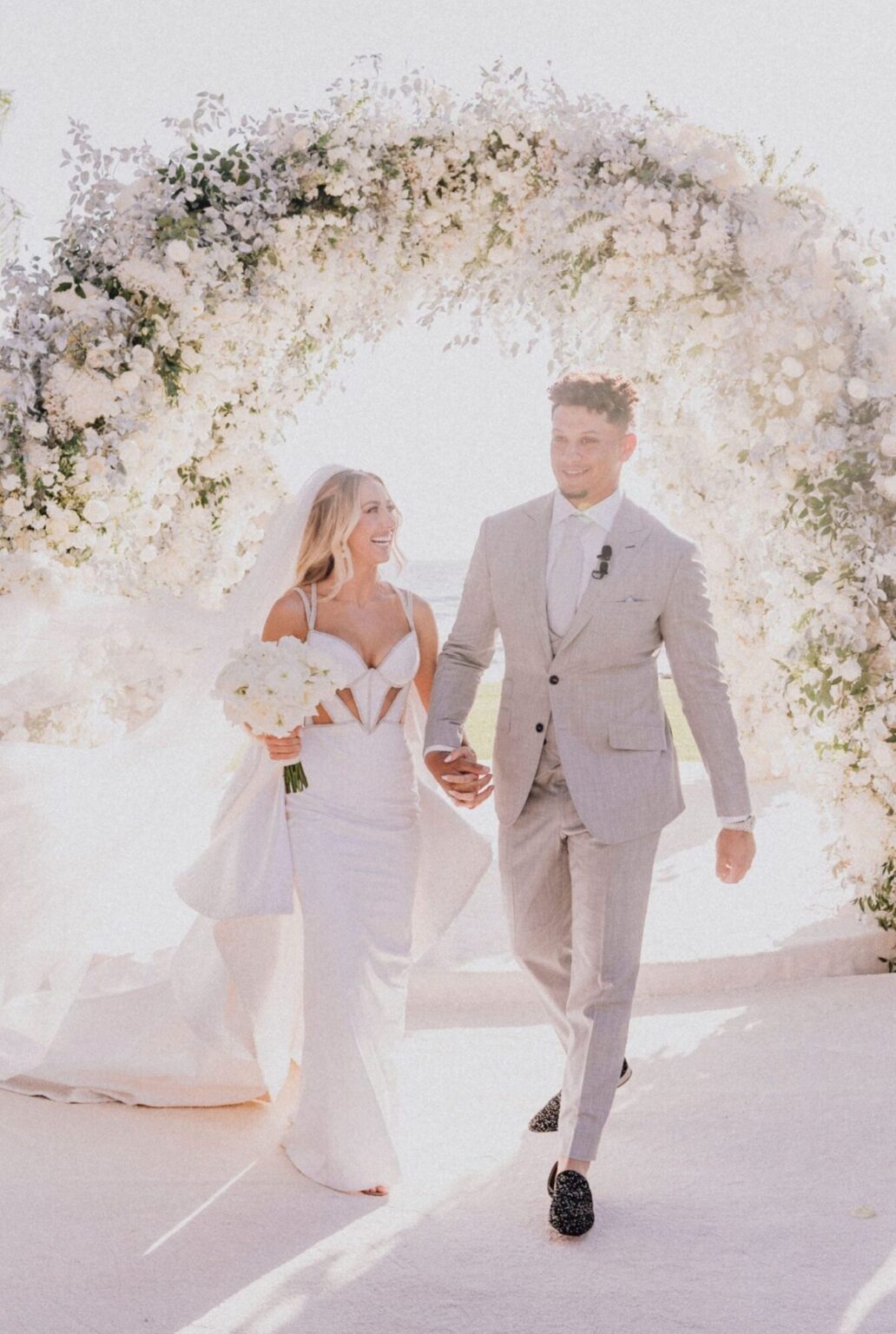 A couple walking hand in hand beneath a large arch of white flowers, Mindy Weiss Party Consultants
