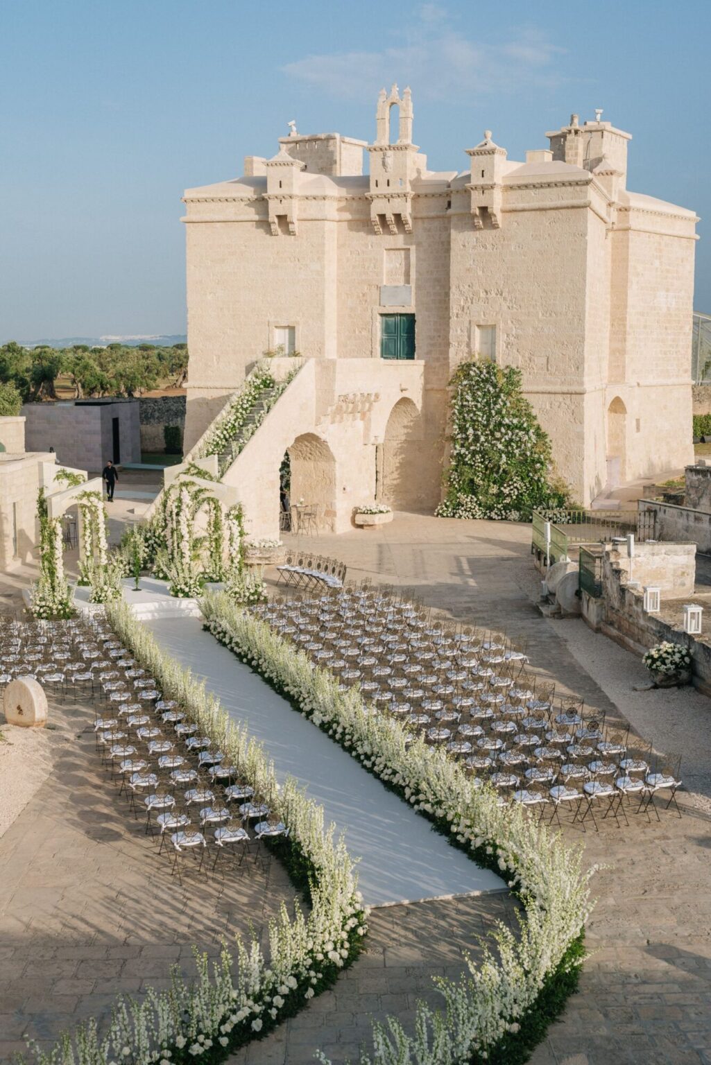 A curving aisle of chairs below a stone castle, its steps edged with white flowers