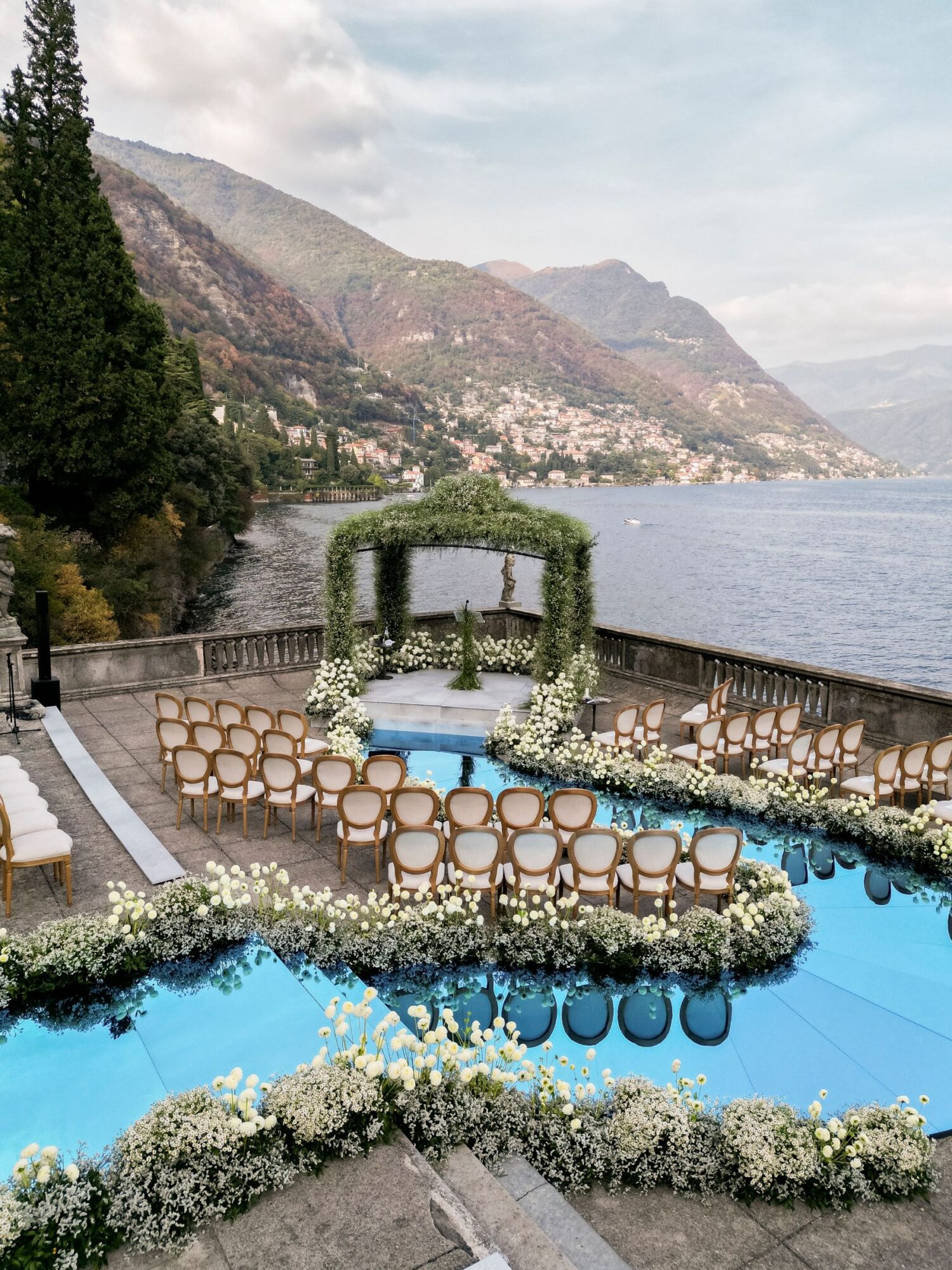 A ceremony laid out beside a pool on a lakeside terrace, with an arch and rows of chairs, Mindy Weiss Party Consultants