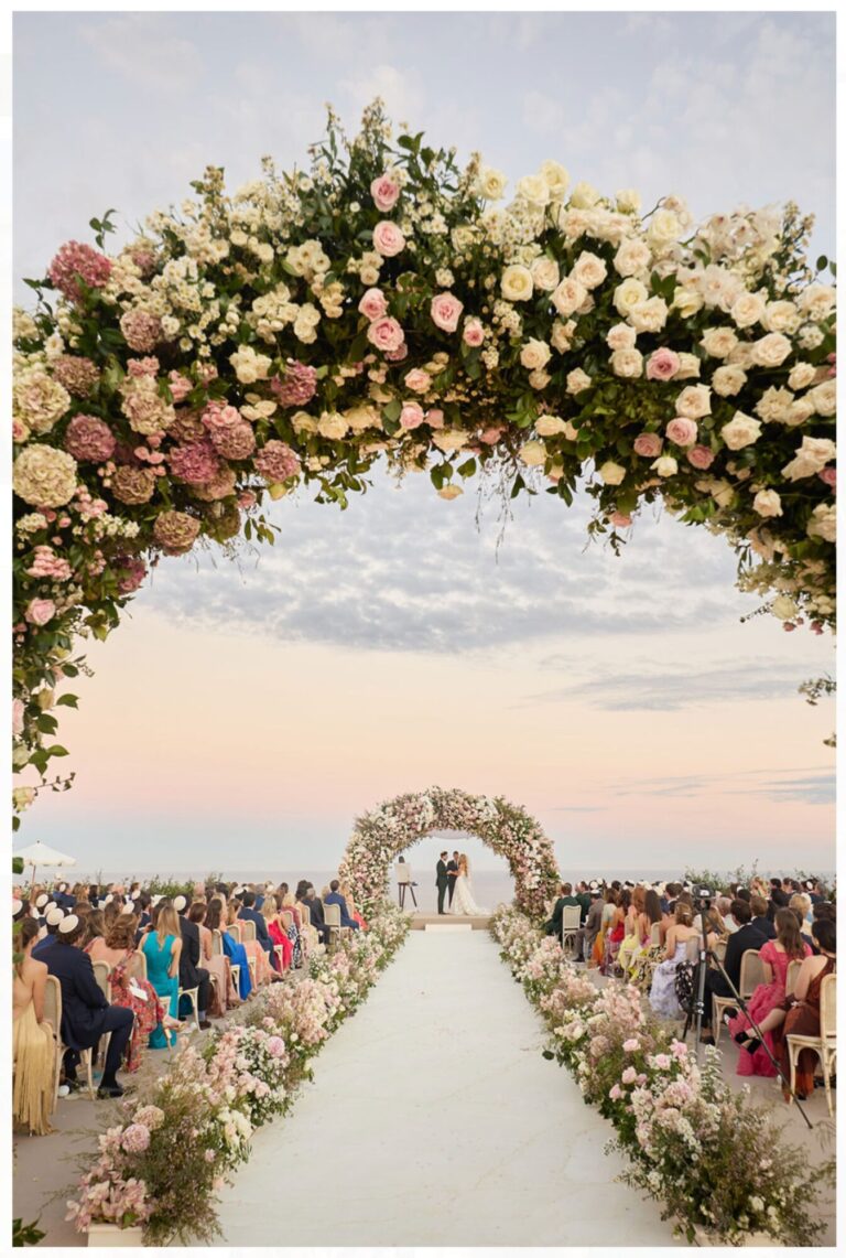 A white aisle edged with flowers leading to a floral arch at sunset, guests seated either side, Mindy Weiss Party Consultants