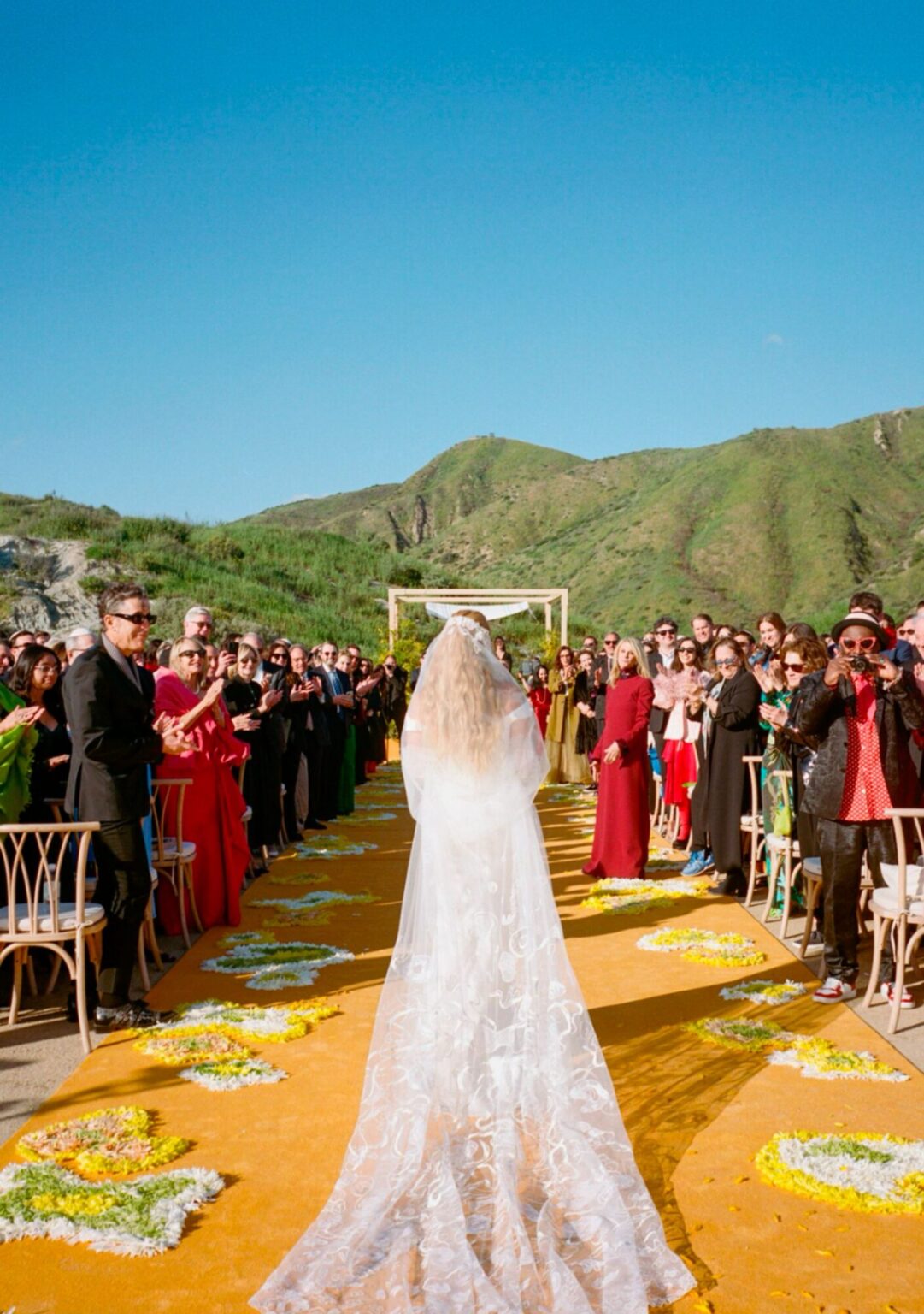 A bride walking a golden-yellow aisle towards guests in the hills, Mindy Weiss Party Consultants