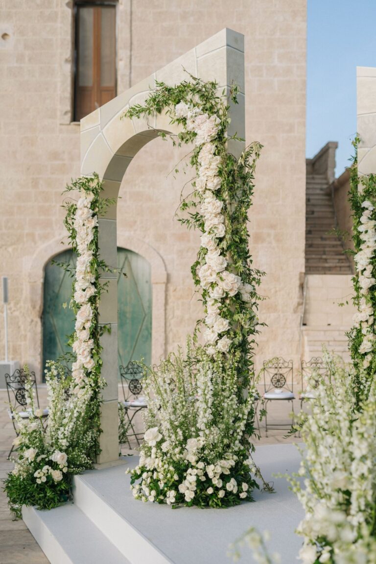 Tall arches wrapped in white flowers and greenery beside a stone building, Mindy Weiss Party Consultants