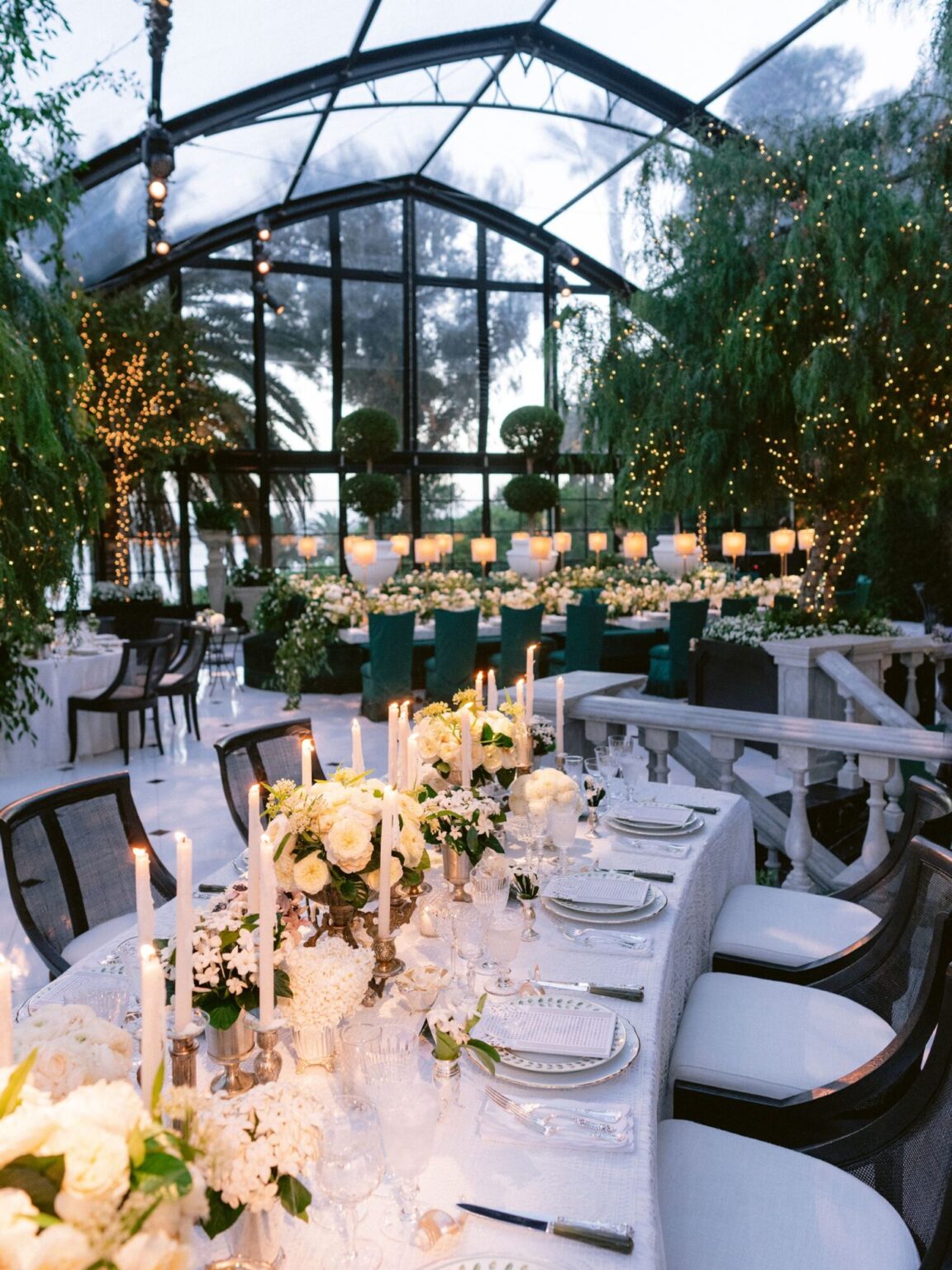 A candlelit long table set with white flowers inside a glass greenhouse