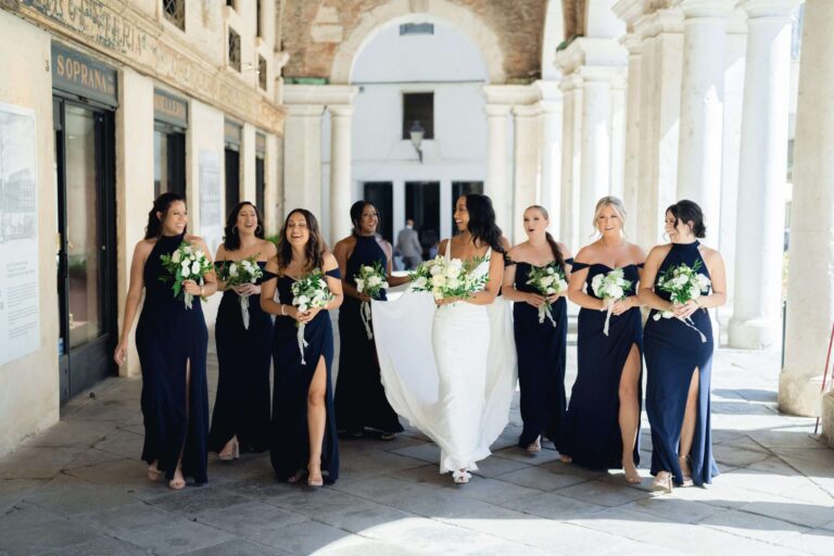 Bride and bridesmaids in navy gowns walking beneath a colonnaded portico, More Than Weddings
