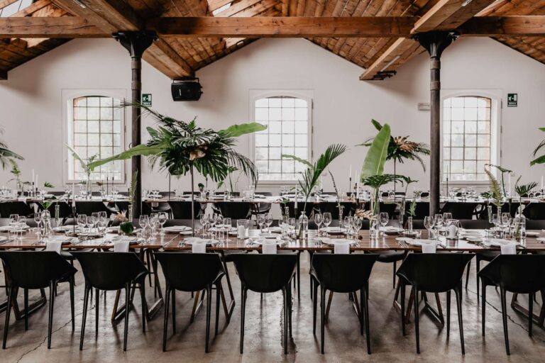Long tables with palm and banana-leaf centrepieces under timber beams in an industrial venue, More Than Weddings