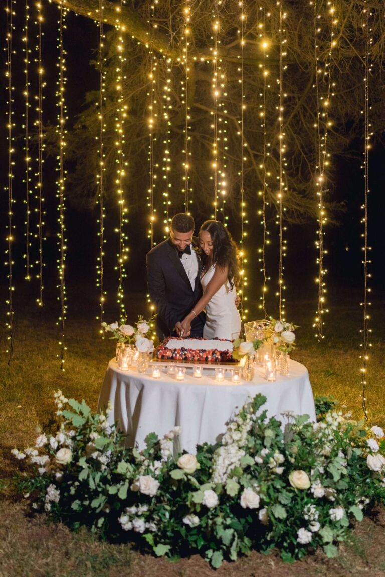 Couple cutting the cake under a curtain of fairy lights, white flowers around the table, More Than Weddings