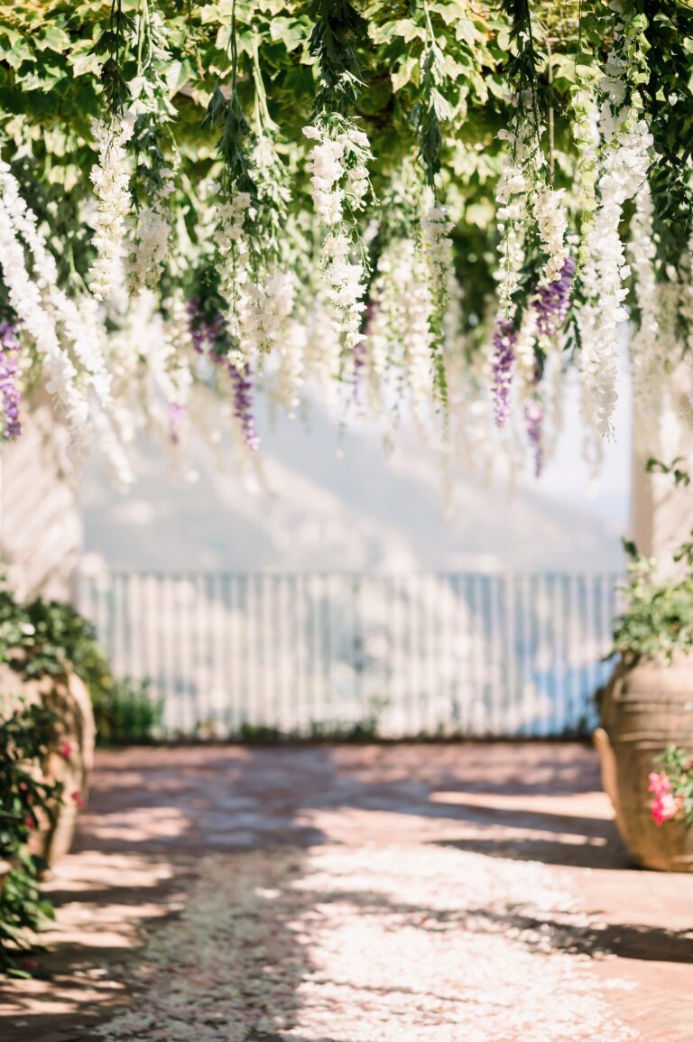 The ceremony aisle under hanging white and lilac blooms, a gate and the sea light at the end of it, Mr and Mrs Wedding In Italy