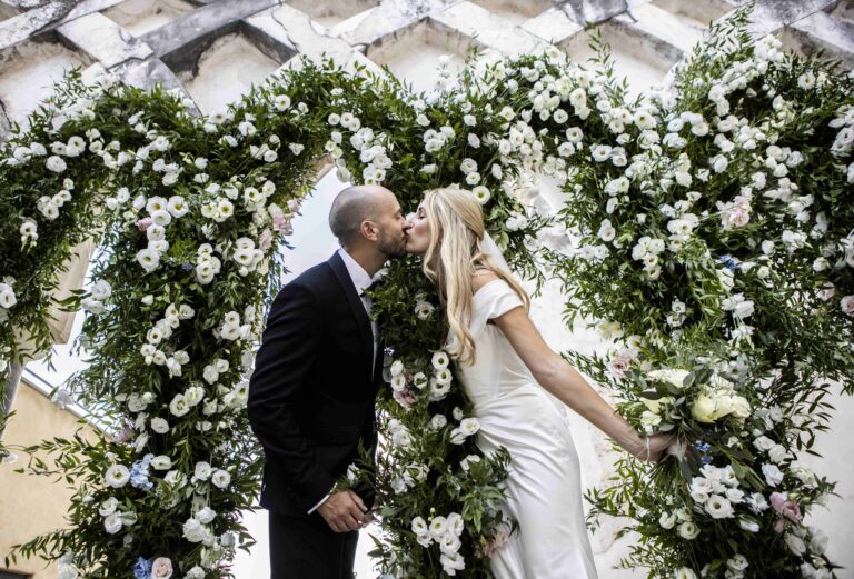 A kiss inside an arch of white flowers and greenery, Puglia, Mr and Mrs Wedding In Italy