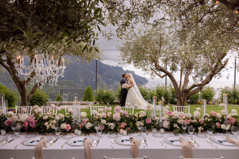 A long table of pink and white peonies laid under olive trees hung with crystal chandeliers, the couple behind it and the Amalfi Coast mountains beyond, Mr and Mrs Wedding In Italy