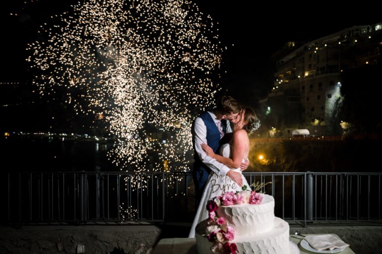 The cake cut under a fountain of fireworks, the bay lit up behind, Mr and Mrs Wedding In Italy