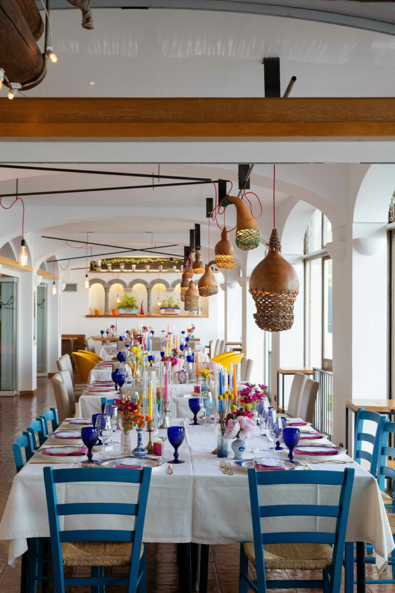 A long table indoors with blue chairs, coloured glassware and hanging lamps, Mr and Mrs Wedding In Italy