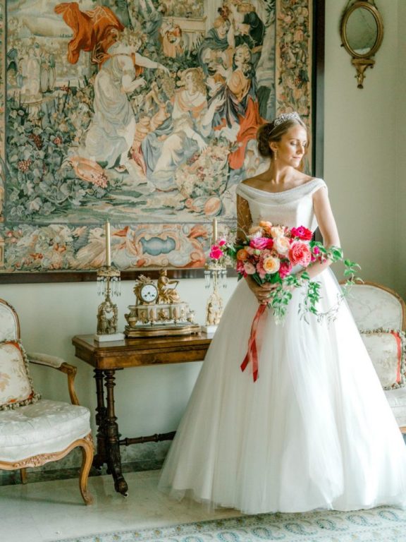 Bride standing beside an antique table with a hand-tied bouquet and red ribbon., My Dubai Wedding Planner