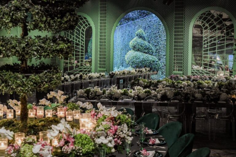 Table of flowers and candles in front of green trellis arches and a lit topiary window, MY Event Design