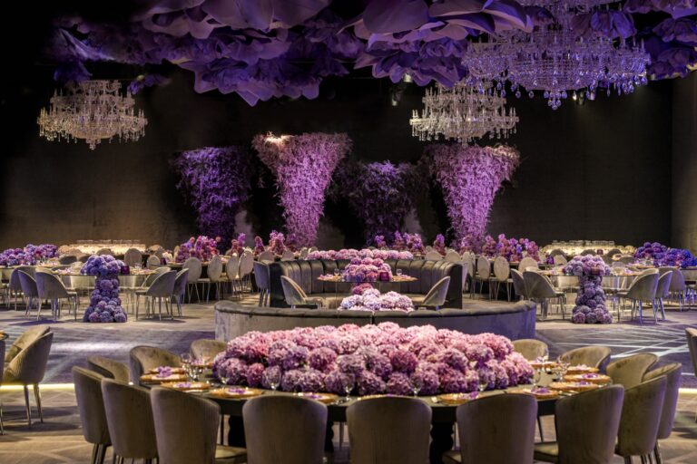 Ballroom with towering purple floral columns, chandeliers and round tables around a central floral display, MY Event Design