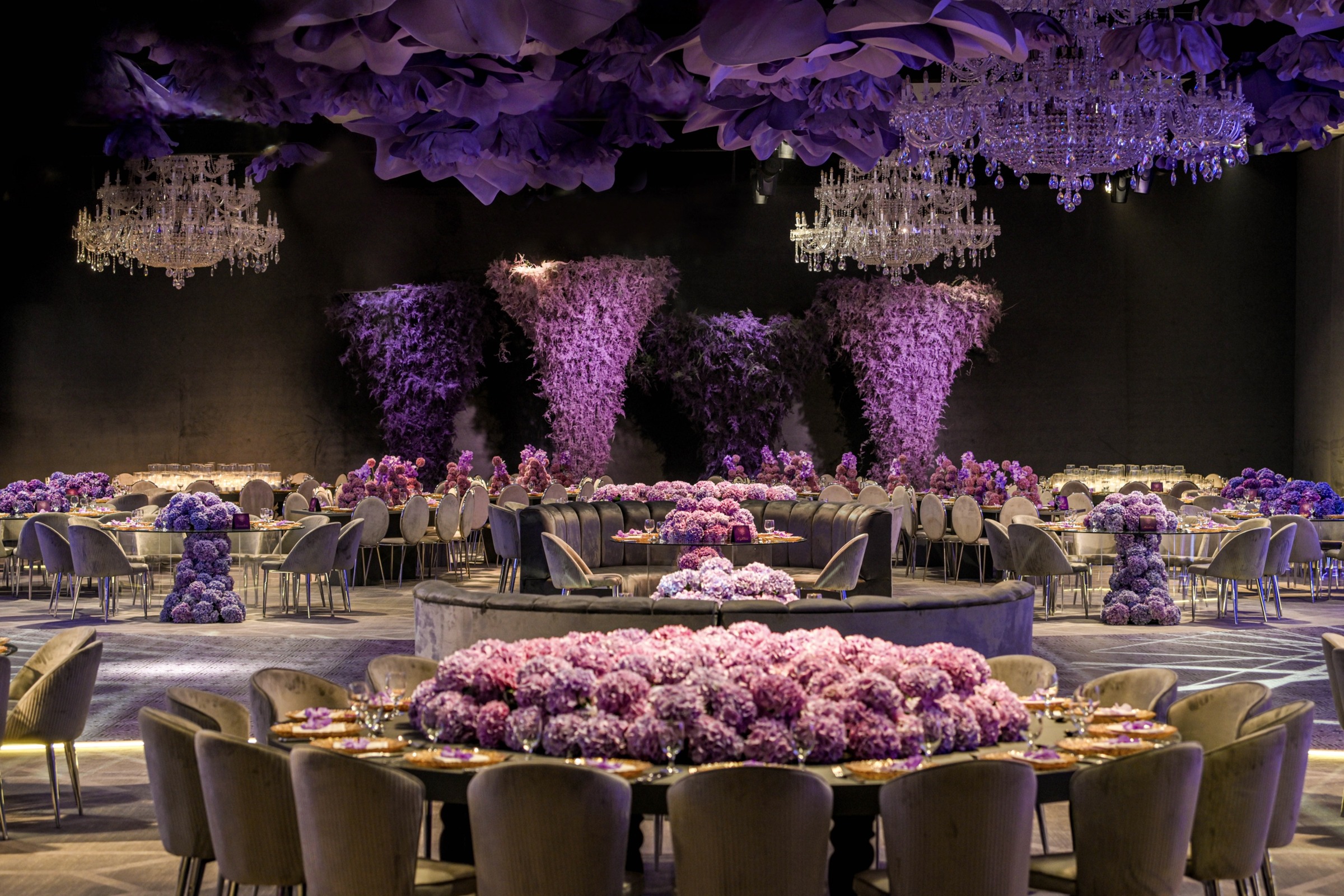 Ballroom with towering purple floral columns, chandeliers and round tables around a central floral display, MY Event Design