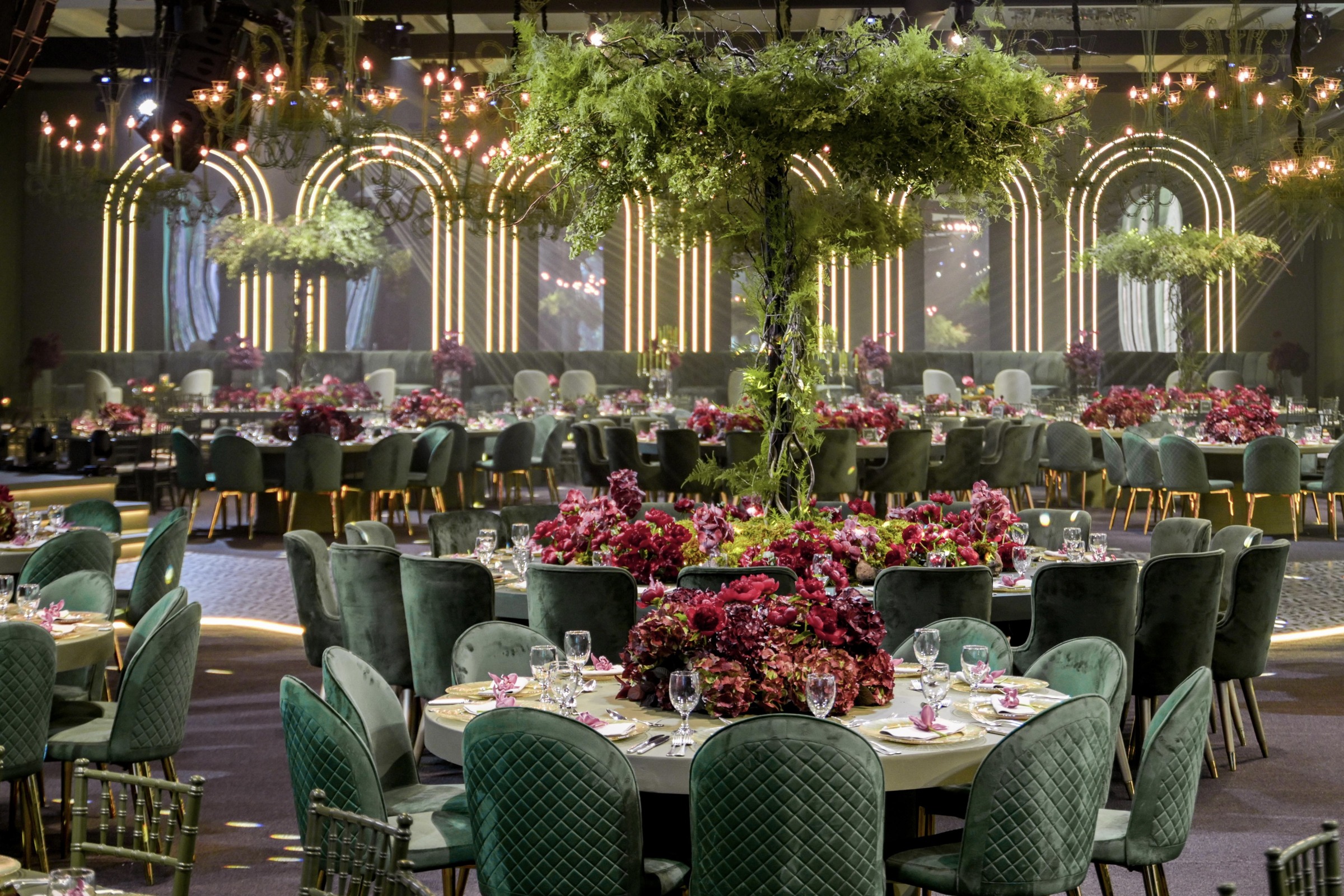 Glasshouse-style hall with illuminated arches, a central living tree and green velvet chairs at dressed tables, MY Event Design